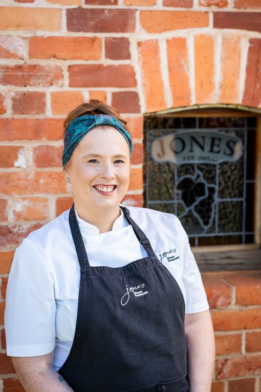 Chef Briony Bradford standing outside Jones Winery Restaurant in Rutherglen, Victoria, wearing a Jones Winery chef uniform in front of the historic red brick winery building and stained glass winery window.