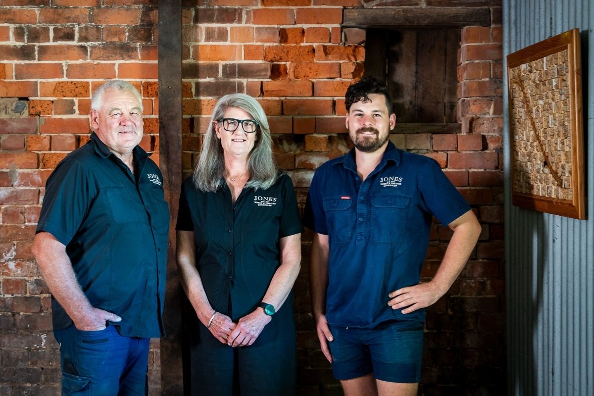 Arthur, Mandy and Ben Jones standing inside the historic brick cellar at Jones Winery & Vineyard in Rutherglen, Victoria. The multi generation family winery owners are dressed in Jones Winery shirts and posed against the original red brick walls of the winery building.